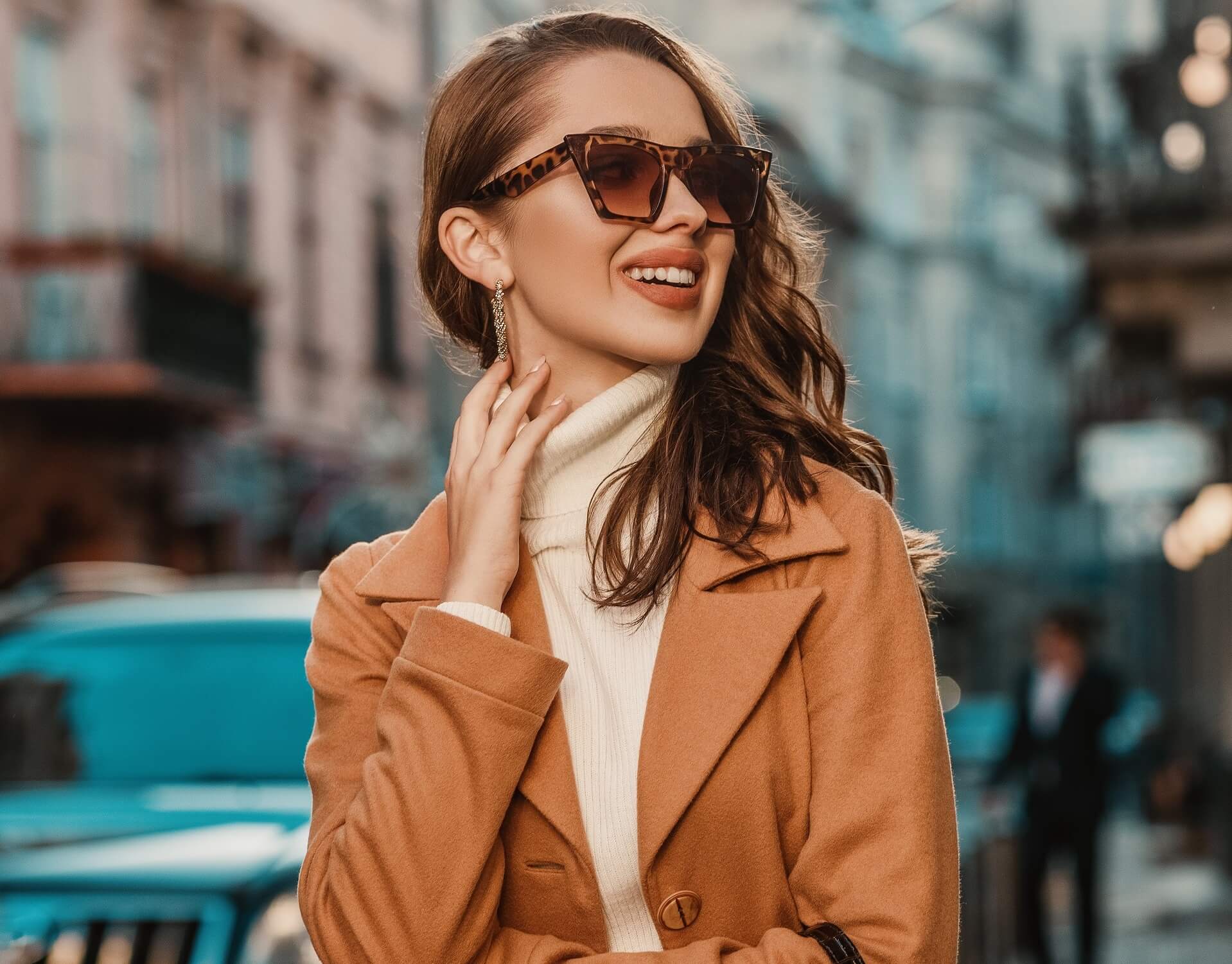 Outdoor portrait of young happy smiling fashionable woman wearing trendy sunglasses, camel color autumn coat, white turtleneck, holding textured reptile handbag, walking in street of European city