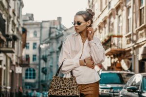 Outdoor autumn fashion portrait of elegant, luxury lady wearing sunglasses, trendy white shirt, wrist watch, holding animal, leopard print bag, posing in street of European city. Copy, empty space