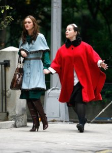 A young glamorous brunette woman walks along a city street in a blue coat, carrying a brown handbag. A middle-aged maid follows close behind in a bright red cape coat.